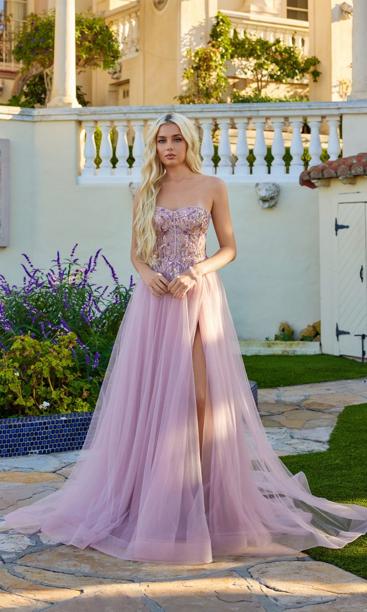 A woman wears the Long Prom Dress: Juliet JT2640G, a strapless light pink tulle gown with a lace-up back and thigh-high slit, standing outdoors on a stone path in front of a building with pillars and a balcony.
